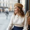 Mujer sonriendo, sentada en un café, mirando por una ventana con vista a la calle. Lleva un sombrero canotier de paja y una blusa blanca con cuello bordado.