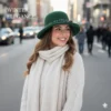 Mujer sonriente en una calle urbana, vestida con un sombrero de fieltro verde botella adornado con hojas y una piña, y un conjunto de suéter de punto trenzado y bufanda largos en color crema.