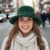 Una mujer joven y sonriente con un sombrero de fieltro verde oscuro, un suéter de punto color crema y una bufanda a juego. Está parada en una calle de la ciudad con autos y peatones borrosos al fondo. El sombrero tiene un adorno de hojas y una pequeña piña. El texto superpuesto en la esquina superior izquierda dice "WINTER ELEGANCE" y un pequeño copo de nieve está en la esquina inferior derecha.