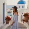 Una mujer con un vestido de rayas, con los hombros al descubierto, y un sombrero fedora negro, está de pie en unas escaleras de piedra blanca en un pueblo de una isla griega, mirando hacia un puerto con barcos y un edificio blanco coronado con una cúpula azul.